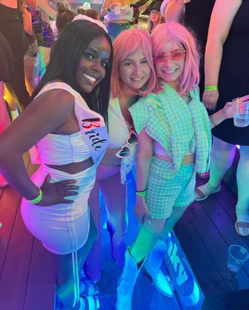 Neon-lit dance floor at a bachelorette party with three friends posing — a smiling woman in a white dress and 'Bride' sash flanked by two pals in pink wigs, colorful outfits and drinks.
