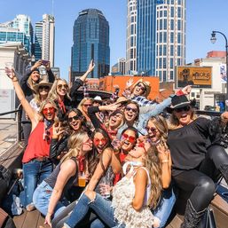 Cheerful group of friends in sunglasses posing with drinks on a sunny downtown rooftop, tall city skyscrapers behind them.