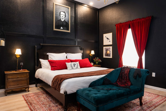 Moody boutique hotel bedroom with black paneled walls, king bed dressed in white linens and red accent pillows, teal velvet chaise, red curtains, wooden nightstands and warm bedside lamps