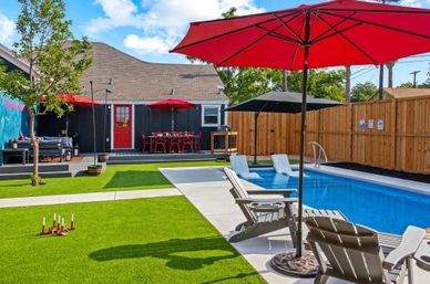 Sunny backyard pool scene with a rectangular blue inground pool, lounge chairs under red umbrellas, artificial turf lawn, deck with red bistro table and seating, and a wooden privacy fence.