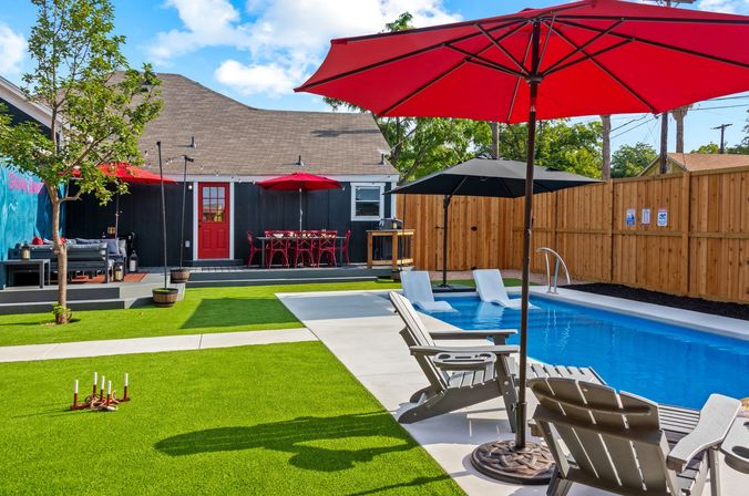 Sunny backyard pool scene with a rectangular blue inground pool, lounge chairs under red umbrellas, artificial turf lawn, deck with red bistro table and seating, and a wooden privacy fence.