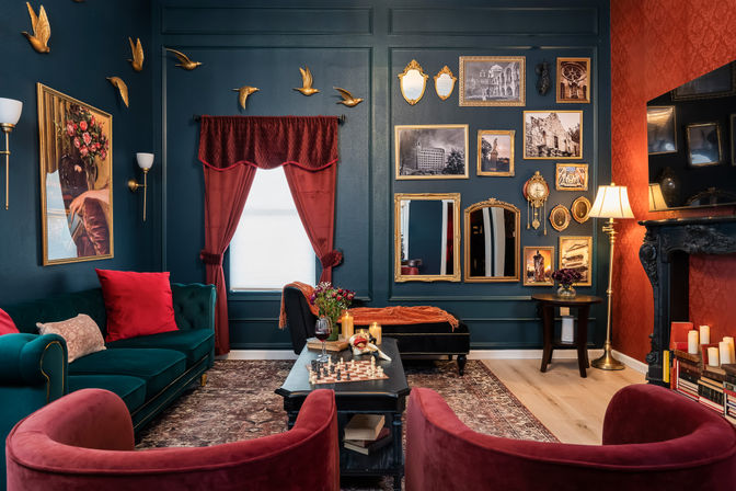 Victorian-style living room interior with dark teal paneled walls, red velvet curtains and chairs, emerald velvet sofa, gold-framed gallery wall and mirrors, decorative brass bird sculptures, ornate black fireplace, coffee table with chessboard and candles, cozy vintage-chic decor.