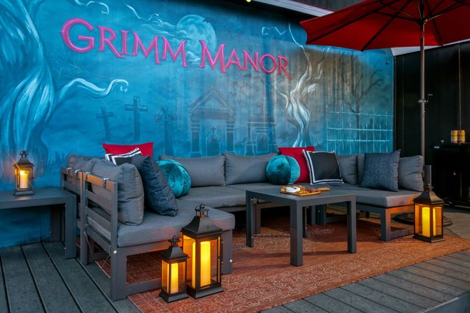 Cozy outdoor patio lounge with gray sectional sofa, teal and red pillows, warm glowing lanterns and a blue spooky mural with pink lettering and graveyard motifs.