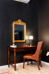 Cozy home-office nook with a vintage dark wood writing desk, brown leather chair, ornate gold-framed mirror, and brass table lamp against a matte black accent wall.