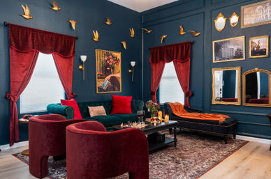 Victorian-inspired living room with dark teal paneled walls, red velvet curtains and chairs, emerald sofa, gold bird wall sculptures, ornate gilt mirrors and patterned rug