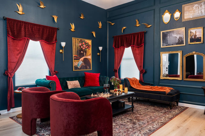 Victorian-inspired living room with dark teal paneled walls, red velvet curtains and chairs, emerald sofa, gold bird wall sculptures, ornate gilt mirrors and patterned rug