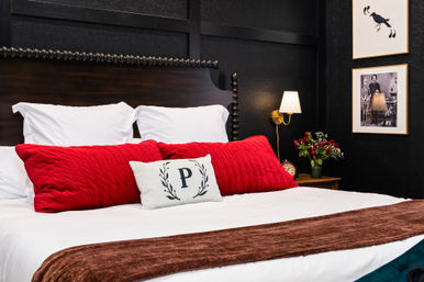 Stylish bedroom with crisp white bedding, pop of red quilted lumbar pillows and a monogram “P” cushion on a dark wood headboard against a black paneled wall, brass wall sconce, bedside bouquet and vintage framed art