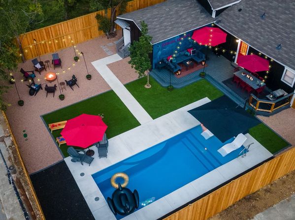 Aerial view of a residential backyard at dusk featuring a rectangular blue pool with a black swan float and yellow ring, white loungers under a dark umbrella, bright red patio umbrellas by seating areas, artificial turf walkways, a deck with string lights and cozy outdoor sofas, and a fire pit surrounded by chairs.
