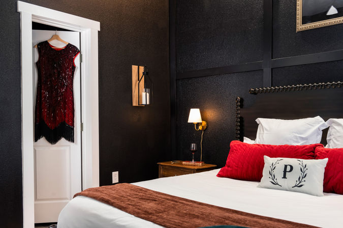 Cozy boutique-style bedroom with black textured walls, king bed dressed in white linens and red pillows, wooden nightstand with lamp and wine glass, and a beaded red flapper dress hanging on a white door.