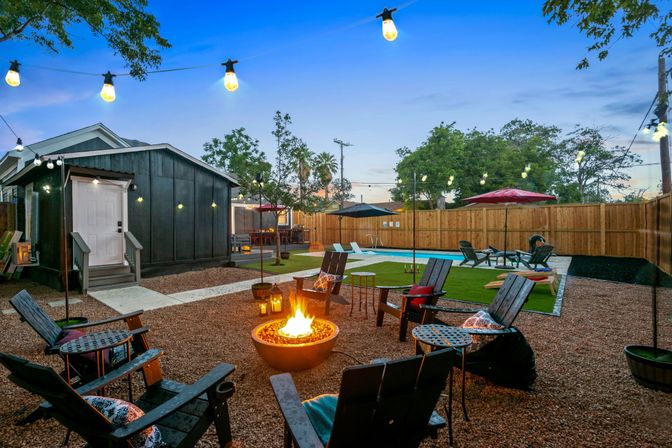 Cozy backyard entertaining space at twilight with a glowing fire pit surrounded by Adirondack chairs, string lights overhead, a black shed, artificial turf and a swimming pool with umbrellas behind a wooden privacy fence.