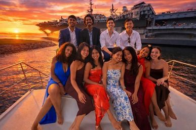 Ten friends in evening attire posing on a yacht during a colorful sunset harbor cruise, with a large aircraft carrier silhouetted against an orange sky.