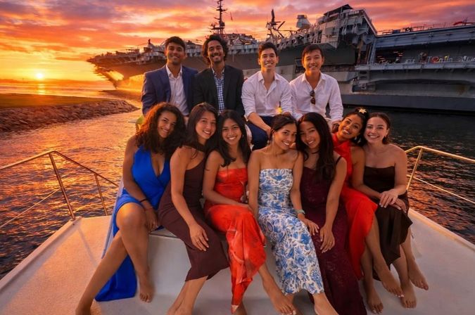 Ten friends in evening attire posing on a yacht during a colorful sunset harbor cruise, with a large aircraft carrier silhouetted against an orange sky.