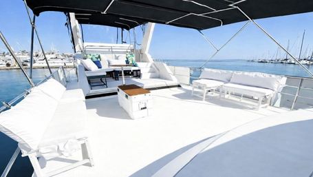 Sun-drenched luxury yacht deck at a marina, featuring spacious white cushioned lounges, a small table and cooler under a black bimini, with calm blue sea and sailboats in the distance