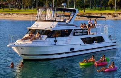 White party yacht anchored in San Diego bay with people sunbathing on the deck and friends relaxing on bright inflatable tubes in calm blue water, tree-lined shoreline in the background.