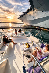People enjoying champagne and snacks on a luxury yacht at golden sunset in a harbor beside a massive moored aircraft carrier