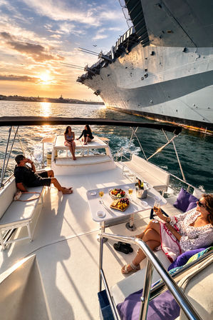 People enjoying champagne and snacks on a luxury yacht at golden sunset in a harbor beside a massive moored aircraft carrier