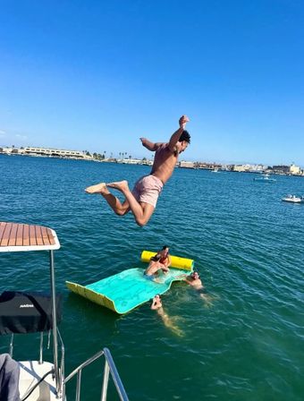 Man mid-air jumping from a boat toward a turquoise inflatable water mat as friends splash in a sunny coastal bay