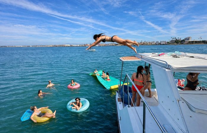 Group enjoying a sunny yacht party in a blue coastal bay — woman mid-dive from boat toward people on colorful inflatable tubes near a distant marina skyline.