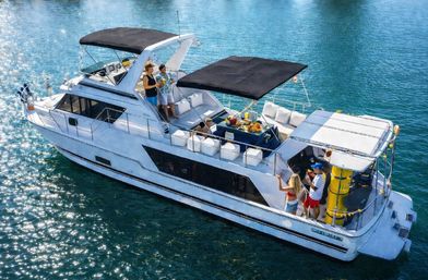 White luxury motor yacht in sparkling blue coastal water, friends socializing on shaded deck with drinks, snacks and black sun canopies