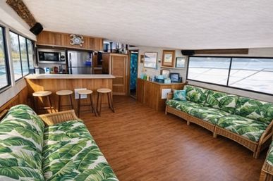 Bright, vacation-ready houseboat interior with wicker sofas in green banana-leaf cushions, wood-look floor, breakfast bar with four stools, stainless steel fridge and microwave, and large windows overlooking the water.