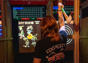 Person seen from behind in an indoor axe‑throwing lane, raising a green‑handled axe toward an electronic target and scoreboard showing cartoon birds and a rubber duck.