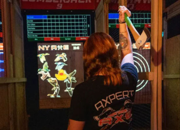 Person seen from behind in an indoor axe‑throwing lane, raising a green‑handled axe toward an electronic target and scoreboard showing cartoon birds and a rubber duck.