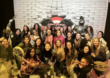 Large group of smiling women playfully posing with foam axes, shields and a costumed knight in front of a white brick backdrop reading “New York” at a themed team-building event.