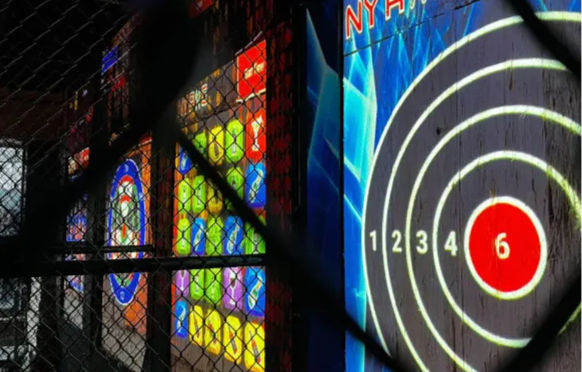 View through a chain-link safety fence of an indoor target lane with an illuminated circular scoring board showing numbered rings and a red bullseye, next to colorful arcade-style light panels.