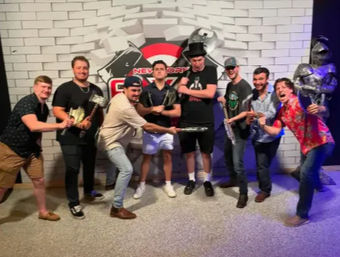 Eight men playfully posing with foam axes, hammers and swords in front of a New York-themed brick photo wall and a suit-of-armor prop at an indoor entertainment venue.