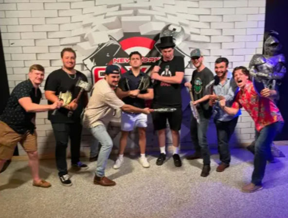 Eight men playfully posing with foam axes, hammers and swords in front of a New York-themed brick photo wall and a suit-of-armor prop at an indoor entertainment venue.