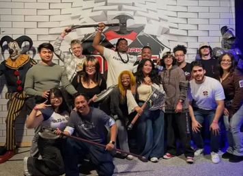 Group of friends posing with oversized battle axes and props at an indoor escape-room attraction, flanked by a costumed jester and armored knight.