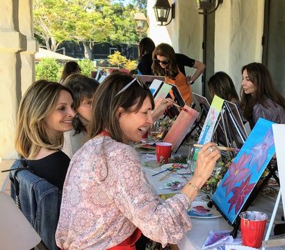 Women enjoying a paint-and-sip art party on a sunlit outdoor patio, smiling and painting colorful floral canvases at a long table with easels, brushes and drinks.