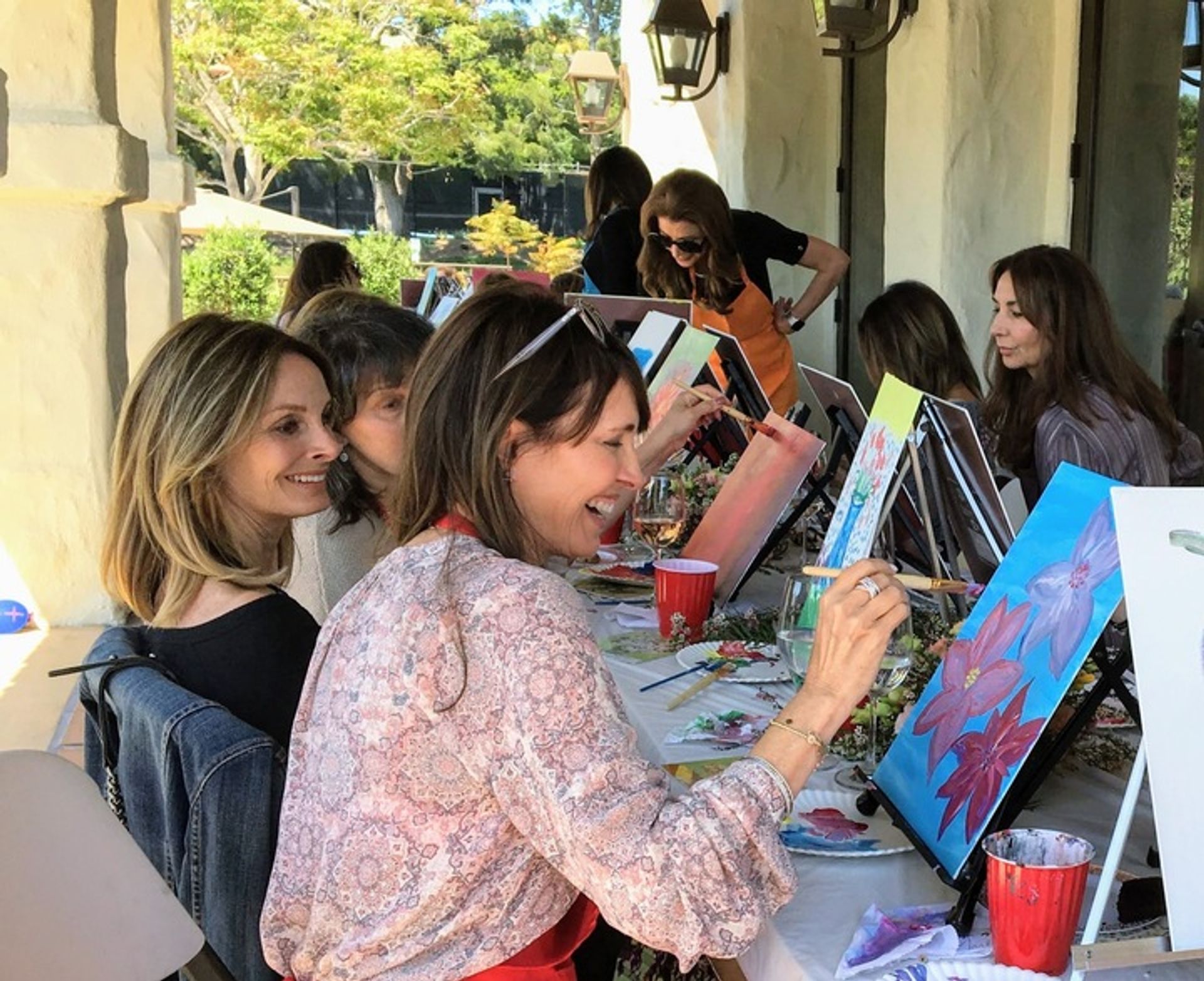 Women enjoying a paint-and-sip art party on a sunlit outdoor patio, smiling and painting colorful floral canvases at a long table with easels, brushes and drinks.