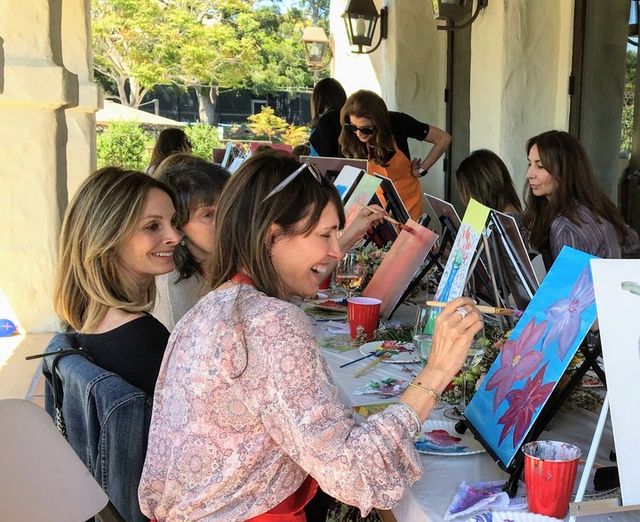 Women enjoying a paint-and-sip art party on a sunlit outdoor patio, smiling and painting colorful floral canvases at a long table with easels, brushes and drinks.