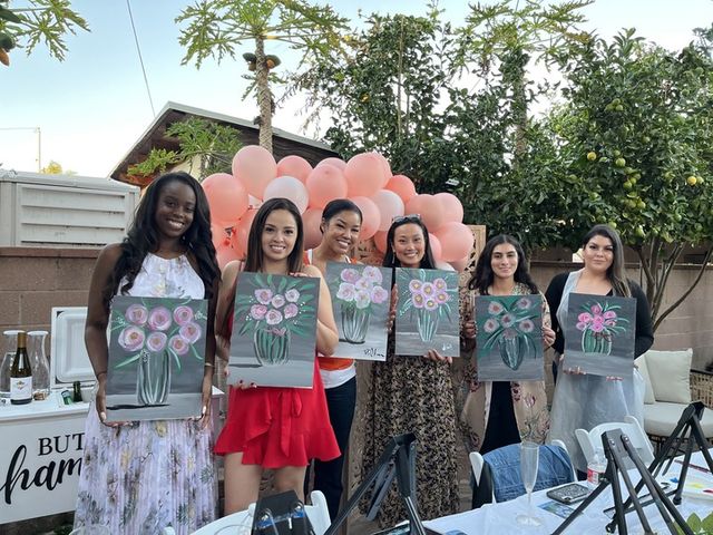 Backyard paint-and-sip: six women hold vase-and-flower canvases in front of a pink balloon arch on an outdoor patio with palm and citrus trees, wine bottles and painting easels on tables.