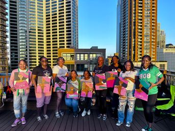 Nine women on an urban rooftop at sunset holding colorful pink-and-green portrait canvases with tall residential skyscrapers in the background — rooftop paint night.