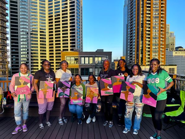 Nine women on an urban rooftop at sunset holding colorful pink-and-green portrait canvases with tall residential skyscrapers in the background — rooftop paint night.