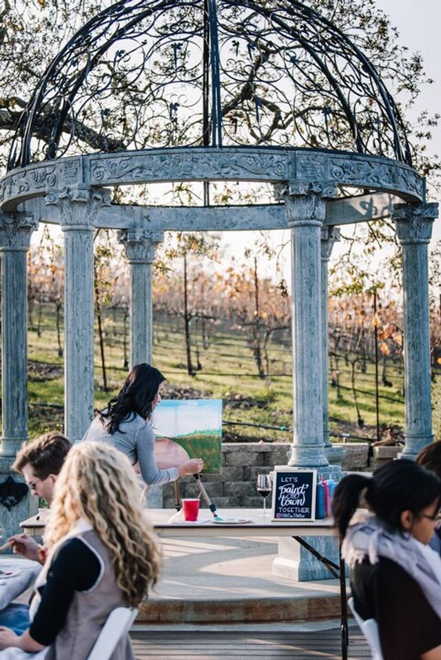 Outdoor painting class under an ornate stone and wrought-iron gazebo, students seated while an instructor works on a vineyard landscape with rows of vines in the background.