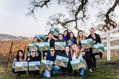 Group of adults at a wine-country vineyard painting party, each holding a vineyard landscape canvas and a glass of wine beneath a gnarled oak and white fence