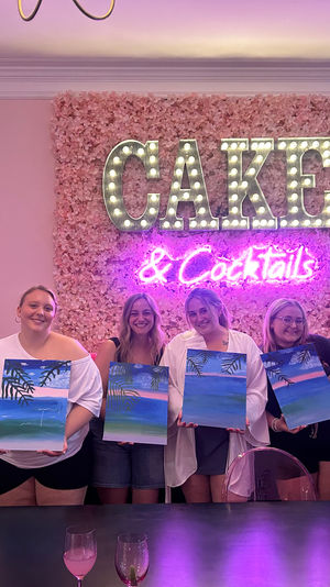 Paint-and-sip party: a group of attendees holding tropical beach canvases in front of a pink floral wall with a lit marquee "CAKE" sign and neon "& Cocktails".