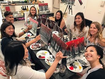 Smiling group at an indoor paint-and-sip party painting matching red autumn-tree canvases around a table with palettes, brushes, cups and wine bottles visible