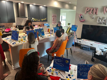 Paint-and-sip party in a modern home kitchen and living area, guests seated around an island painting blue night-sky canvases with palettes, drinks, a "But first mimosas" banner and pink balloons.