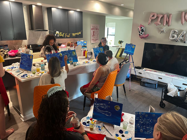 Paint-and-sip party in a modern home kitchen and living area, guests seated around an island painting blue night-sky canvases with palettes, drinks, a "But first mimosas" banner and pink balloons.