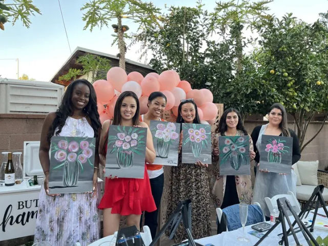 Six women at an outdoor backyard painting party holding matching floral canvas paintings in front of a pink balloon garland, with easels, drinks and patio greenery visible.
