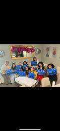 Group of friends at a cozy living-room paint night, smiling and holding blue starry-night canvases under pink "LET'S GO GIRLS" balloon letters