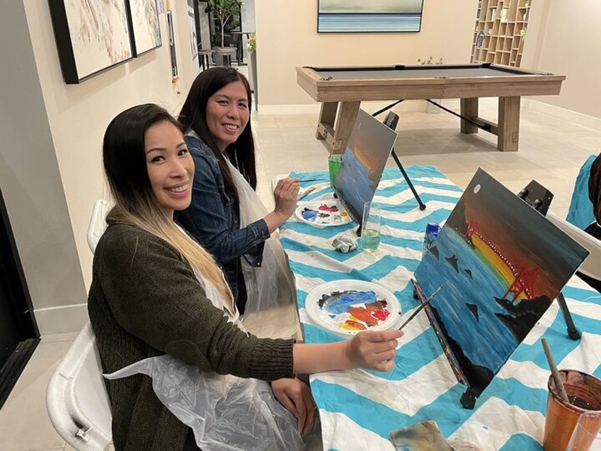 Two people at a paint night table creating sunset bridge canvases on tabletop easels, with paint palettes, brushes and a striped tablecloth in a bright indoor recreation room with a pool table in the background.