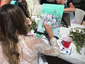 Participant in a lace top painting pink roses on a turquoise canvas at an outdoor paint-and-sip party, table with art supplies, flowers and a wine glass