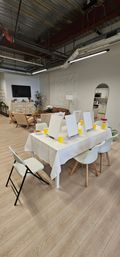 Open-concept modern loft art studio with communal table of easels and yellow cups, cozy lounge seating and visible kitchenette.