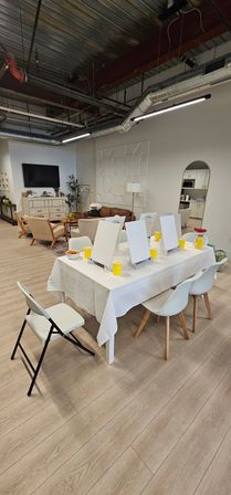 Open-concept modern loft art studio with communal table of easels and yellow cups, cozy lounge seating and visible kitchenette.