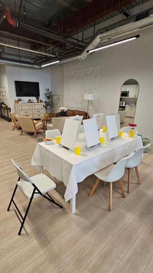 Open-concept modern loft art studio with communal table of easels and yellow cups, cozy lounge seating and visible kitchenette.